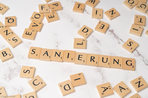 Scrabble-Steine mit dem Wort „Sanierung“ auf Marmor, umgeben von verstreuten Buchstaben