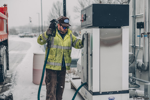 Ein Mann in einer Jacke tankt im Winter bei Schneefall an einer Tankstelle einen Lkw mit Diesel auf.